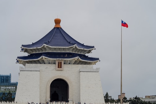 A large, traditional Chinese-style building with a dark blue tiled roof and white walls stands prominently. The structure features tiered eaves with intricate details, and an arched entrance is visible. In the background, a tall flagpole with a red flag flies against a cloudy sky. A group of people can be seen near the base of the building.
