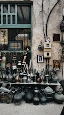 A collection of vintage metal jugs and pots is displayed on shelves against a rustic stone wall. Decorative items hang on the wall, including framed art and Arabic calligraphy. The setting has an old-world charm, highlighted by the weathered textures and patina on the items.