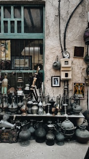 A collection of vintage metal jugs and pots is displayed on shelves against a rustic stone wall. Decorative items hang on the wall, including framed art and Arabic calligraphy. The setting has an old-world charm, highlighted by the weathered textures and patina on the items.