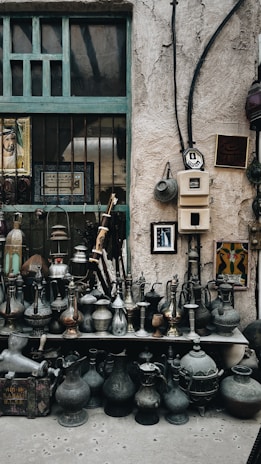 A collection of vintage metal jugs and pots is displayed on shelves against a rustic stone wall. Decorative items hang on the wall, including framed art and Arabic calligraphy. The setting has an old-world charm, highlighted by the weathered textures and patina on the items.