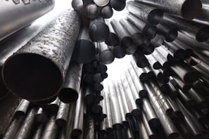 A collection of large, metallic tubes of varying sizes arranged in a chaotic yet artistic pattern. The tubes are seen from a low angle, looking upwards, with a focus on their circular openings and the texture of their surfaces.
