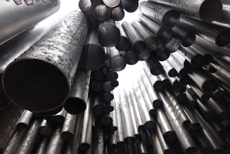 A collection of large, metallic tubes of varying sizes arranged in a chaotic yet artistic pattern. The tubes are seen from a low angle, looking upwards, with a focus on their circular openings and the texture of their surfaces.
