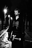 A person in traditional martial arts attire, holding a shinai, stands on a cobblestone path lit by street lamps. The scene is in black and white with the path and lamps creating a dramatic, shadowed effect.
