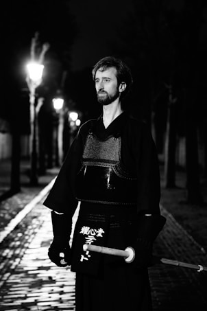 A person in traditional martial arts attire, holding a shinai, stands on a cobblestone path lit by street lamps. The scene is in black and white with the path and lamps creating a dramatic, shadowed effect.