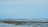 A shoreline with shallow water and a sandy beach, where several cargo ships are visible on the distant horizon. One person is walking along the water’s edge.