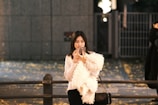 A woman with long hair is focused on her smartphone. She is wearing a fluffy white coat and standing outdoors in front of a large gate. The ground is covered with scattered yellow leaves, suggesting an autumn setting. The scene is dimly lit with artificial lighting, creating a cozy night-time atmosphere.