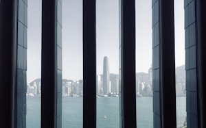 Tall vertical windows frame a panoramic view of a modern cityscape with a distinctive, tall skyscraper at the center. The buildings are set against a clear sky and a body of water in the foreground, enhancing the urban feel.