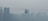 A cable car is suspended across a skyline dominated by tall, barely visible skyscrapers obscured by heavy smog. The muted tones and blurred outlines suggest a setting with high levels of air pollution.