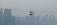 A cable car is suspended across a skyline dominated by tall, barely visible skyscrapers obscured by heavy smog. The muted tones and blurred outlines suggest a setting with high levels of air pollution.