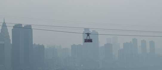 A cable car is suspended across a skyline dominated by tall, barely visible skyscrapers obscured by heavy smog. The muted tones and blurred outlines suggest a setting with high levels of air pollution.