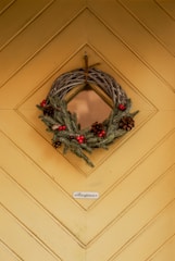 A decorative wreath hangs on an intricately designed wooden door. The wreath is made of twigs, evergreen branches, pine cones, and red berries. Below the wreath, there is a small sign that reads 'Reception'. The wood is painted in a warm, muted yellow and features a chevron pattern.