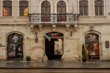 A historic building with arched windows and an ornate balcony adorned with decorative greenery. The facade is decorated with flags and there are two storefronts on either side of a central archway, one displaying a colorful interior. The street in front is wet, likely from rain, and a small pile of sand or debris rests on the pavement.
