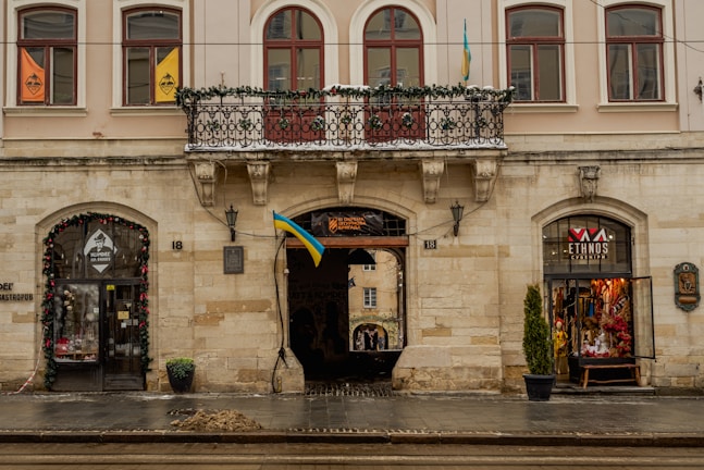 A historic building with arched windows and an ornate balcony adorned with decorative greenery. The facade is decorated with flags and there are two storefronts on either side of a central archway, one displaying a colorful interior. The street in front is wet, likely from rain, and a small pile of sand or debris rests on the pavement.