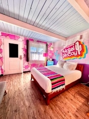 A vibrant hotel room featuring a colorful mural on the wall with the text 'Tucson Hotel McCoy.' The room includes a large bed with a decorative striped blanket, wooden flooring, and a pink lampshade. Pink geometric patterns adorn the wall, along with a blue ceiling and a door with a window.