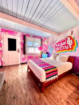 A vibrant hotel room featuring a colorful mural on the wall with the text 'Tucson Hotel McCoy.' The room includes a large bed with a decorative striped blanket, wooden flooring, and a pink lampshade. Pink geometric patterns adorn the wall, along with a blue ceiling and a door with a window.