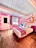 A vibrant hotel room featuring a colorful mural on the wall with the text 'Tucson Hotel McCoy.' The room includes a large bed with a decorative striped blanket, wooden flooring, and a pink lampshade. Pink geometric patterns adorn the wall, along with a blue ceiling and a door with a window.