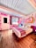 A vibrant hotel room featuring a colorful mural on the wall with the text 'Tucson Hotel McCoy.' The room includes a large bed with a decorative striped blanket, wooden flooring, and a pink lampshade. Pink geometric patterns adorn the wall, along with a blue ceiling and a door with a window.