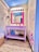 A brightly lit bathroom featuring a pink framed mirror above a lavender sink. The walls are adorned with playful, colorful illustrations, including a purse and other whimsical designs. The floor is a light wood texture, and a wicker basket sits on a lavender shelf. A round magnifying mirror is mounted on the wall.