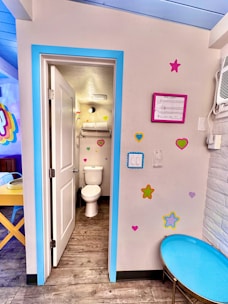 The image depicts a small bathroom area with bright and playful decor. The room has a wooden floor and a white door leading to a toilet area framed with a vibrant blue border. The walls are adorned with colorful heart and star stickers. A blue circular table is seen to the right. Above the toilet, there is a shelf with towels. To the right, an air conditioning unit is visible.
