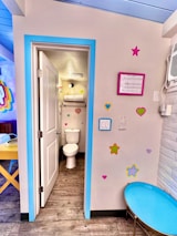 The image depicts a small bathroom area with bright and playful decor. The room has a wooden floor and a white door leading to a toilet area framed with a vibrant blue border. The walls are adorned with colorful heart and star stickers. A blue circular table is seen to the right. Above the toilet, there is a shelf with towels. To the right, an air conditioning unit is visible.