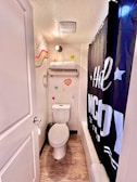 A small bathroom with a toilet, shower, and a wall-mounted towel rack. The walls feature colorful heart decorations and a wavy pattern. The shower curtain is black with bold white text. The floor is textured wood, and the lighting is bright.