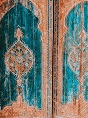 Intricate wooden doors feature elaborate carvings with a symmetrical design, primarily in shades of teal and orange. The decorative pattern includes floral motifs and geometric shapes, set against a richly textured background that shows signs of age and wear.