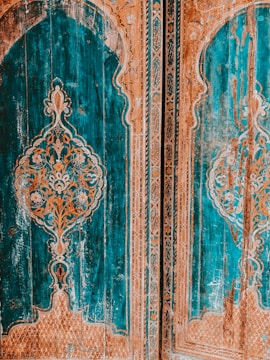 Intricate wooden doors feature elaborate carvings with a symmetrical design, primarily in shades of teal and orange. The decorative pattern includes floral motifs and geometric shapes, set against a richly textured background that shows signs of age and wear.