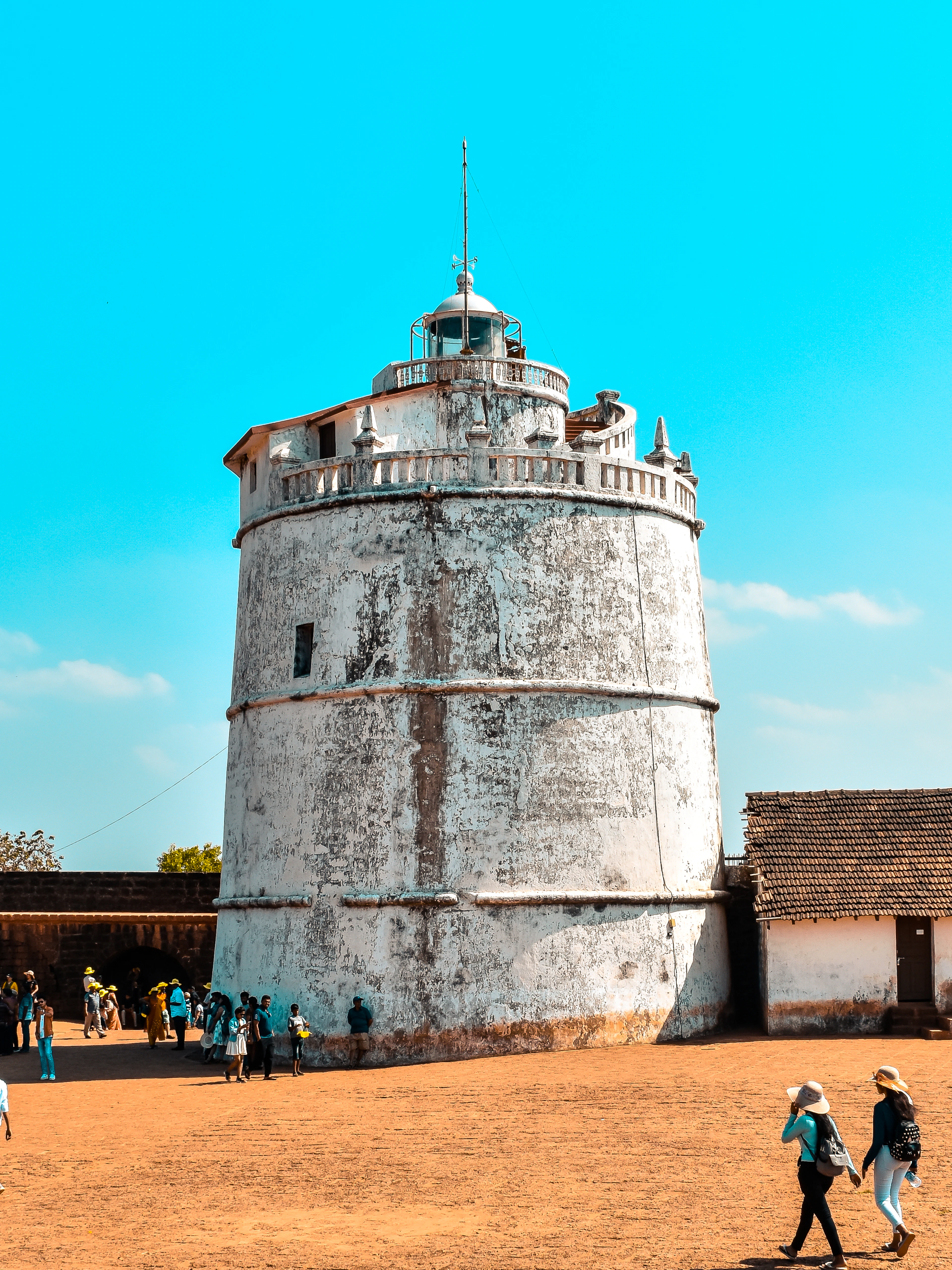 The whitewashed lighthouse tower of Fort Aguada standing on the fort's red laterite ground above Sinquerim, weathered and squat against a vivid blue sky, with visitors crossing the open square below.