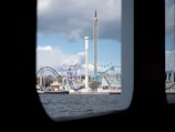 A view of an amusement park seen through the window of a boat. The park features multiple rides including roller coasters and a tall drop tower against a backdrop of partly cloudy skies.