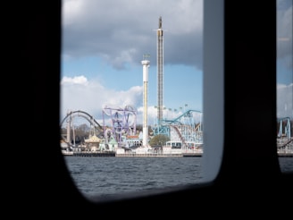 A view of an amusement park seen through the window of a boat. The park features multiple rides including roller coasters and a tall drop tower against a backdrop of partly cloudy skies.