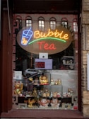 A shop window featuring a colorful neon sign advertising bubble tea, with lively neon colors spelling out 'Bubble Tea'. Below the sign, shelves display a variety of metallic and ceramic cat figurines, commonly known as Maneki-neko, which are popular as good luck charms. Reflections of the city street and buildings can be seen on the glass.