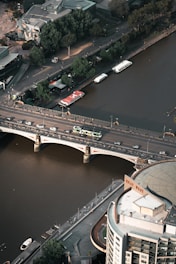 An aerial view of a cityscape featuring a bridge spanning over a river with a tram traveling across. On one side of the river, there are trees lining a pathway with several parked boats along the water’s edge. The opposite side showcases urban architecture, including a prominent building with a curved facade marked 'Quay West.'