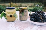Two jars of preserved food with green leaves attached are placed on a lace tablecloth outdoors. One jar has a blue lid and the other has a gold lid. Next to them is a white bowl filled with a large bunch of dark grapes. The background consists of a blurred garden scene.