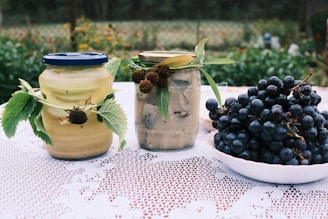 Two jars of preserved food with green leaves attached are placed on a lace tablecloth outdoors. One jar has a blue lid and the other has a gold lid. Next to them is a white bowl filled with a large bunch of dark grapes. The background consists of a blurred garden scene.