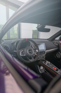 The photograph captures the interior view of a modern car from the driver's seat perspective. The focus is on the sleek steering wheel, dashboard, and control panel, accented with advanced technology and design elements. Outside the car, a glass window and part of a building are visible, suggesting an urban setting.
