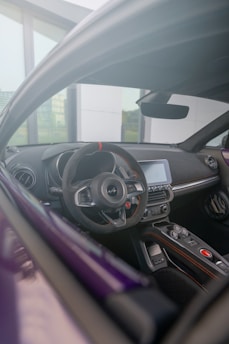 The photograph captures the interior view of a modern car from the driver's seat perspective. The focus is on the sleek steering wheel, dashboard, and control panel, accented with advanced technology and design elements. Outside the car, a glass window and part of a building are visible, suggesting an urban setting.
