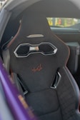 Close-up view of a car seat with a modern, sporty design. The seat is predominantly black with orange stitching and features a metallic handle near the headrest. The car's interior is partially visible with a purple-colored exterior frame in the foreground.
