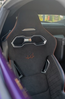 Close-up view of a car seat with a modern, sporty design. The seat is predominantly black with orange stitching and features a metallic handle near the headrest. The car's interior is partially visible with a purple-colored exterior frame in the foreground.