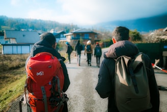 A group of people walking down a paved road in a rural area, carrying backpacks. They are surrounded by houses and open fields, with a backdrop of hills or mountains partially covered by clouds.