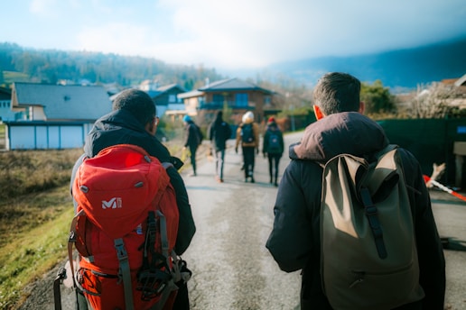 A group of people walking down a paved road in a rural area, carrying backpacks. They are surrounded by houses and open fields, with a backdrop of hills or mountains partially covered by clouds.
