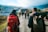 A group of people walking down a paved road in a rural area, carrying backpacks. They are surrounded by houses and open fields, with a backdrop of hills or mountains partially covered by clouds.