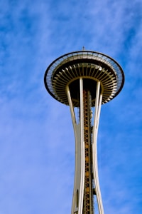 The image features the top of the Space Needle against a clear blue sky. The structure showcases its distinctive saucer-shaped upper section with an observation deck.