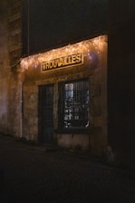 A dimly lit storefront with a rustic appearance is adorned with string lights. The sign above the door displays the word 'Trouvailles'. The cobblestone street leading to the entrance adds an old-world charm.
