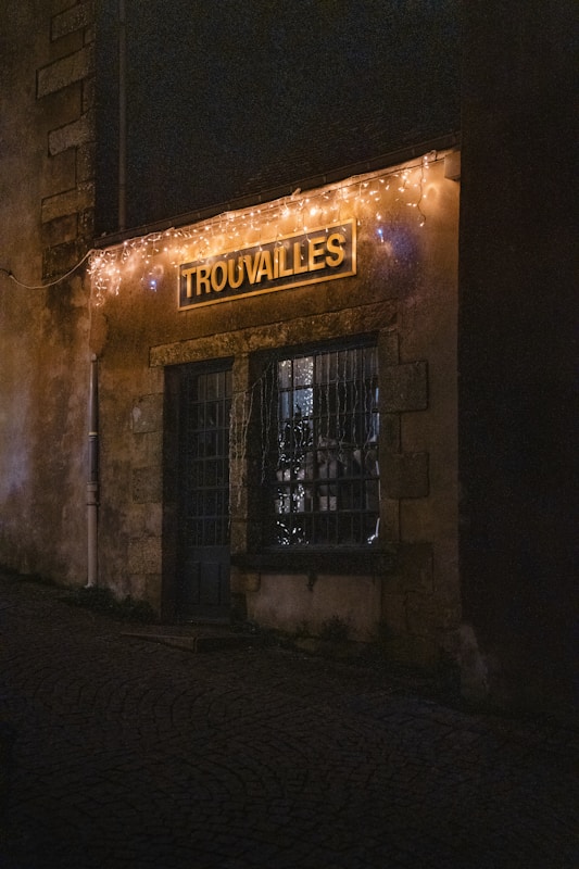 A dimly lit storefront with a rustic appearance is adorned with string lights. The sign above the door displays the word 'Trouvailles'. The cobblestone street leading to the entrance adds an old-world charm.