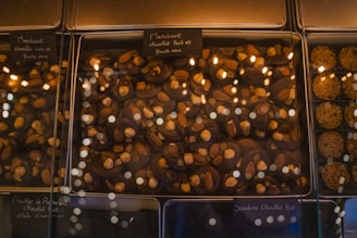 A glass display case presents an assortment of chocolate confections. The chocolates are topped with various nuts, including almonds and hazelnuts. Labels, written in French, are placed above each section, indicating the type of chocolate and toppings. The warm lighting reflects off the glass, adding a cozy and inviting atmosphere.