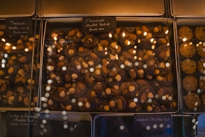 A glass display case presents an assortment of chocolate confections. The chocolates are topped with various nuts, including almonds and hazelnuts. Labels, written in French, are placed above each section, indicating the type of chocolate and toppings. The warm lighting reflects off the glass, adding a cozy and inviting atmosphere.