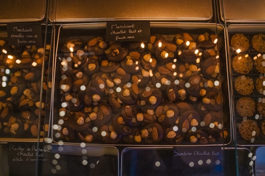 A glass display case presents an assortment of chocolate confections. The chocolates are topped with various nuts, including almonds and hazelnuts. Labels, written in French, are placed above each section, indicating the type of chocolate and toppings. The warm lighting reflects off the glass, adding a cozy and inviting atmosphere.