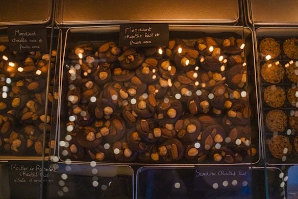 A glass display case presents an assortment of chocolate confections. The chocolates are topped with various nuts, including almonds and hazelnuts. Labels, written in French, are placed above each section, indicating the type of chocolate and toppings. The warm lighting reflects off the glass, adding a cozy and inviting atmosphere.
