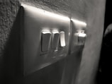 A close-up view of a wall-mounted electrical switch panel with three white switches and a dimly lit background. The surrounding wall has a slightly rough texture, and the scene is captured in grayscale, giving it a vintage feel.