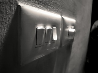 A close-up view of a wall-mounted electrical switch panel with three white switches and a dimly lit background. The surrounding wall has a slightly rough texture, and the scene is captured in grayscale, giving it a vintage feel.